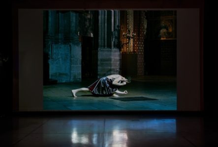 Une salle dans laquelle est projeté le film Transept de Boris Charmatz & César Vayssié. La projection occupe toute la surface du mur. On voit sur l'image un homme, qui porte une jupe. Il effectue un mouvement pour lequel son torse se rapproche du sol. on le voit de profil dans un espace qui pourrait être celui d'une cathédrale.