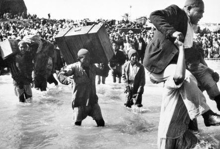 Image en noir et blanc montrant des réfugiés portant des valises et traversant l'eau, entourés d'une foule dense, illustrant l'exode et les défis humanitaires.