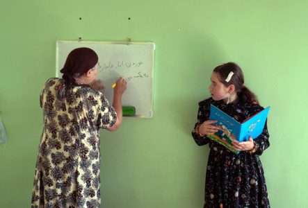 Une femme écrit en langue locale sur un tableau blanc pendant qu'une jeune fille tient un livre coloré, illustrant l'éducation et la transmission du savoir dans un environnement modeste.